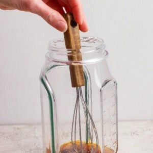 A hand holding a whisk is mixing sauce ingredients in a clear glass jar on a light surface.