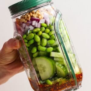 A hand holds a tilted plastic jar filled with layered ingredients including cucumber, edamame, red onion, walnuts, and dressing at the bottom.
