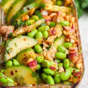 A close-up of a salad with sliced cucumbers, edamame, red onion, chopped nuts, and a red chili-flake dressing in a rectangular glass container.