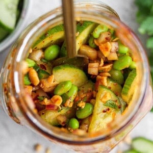 Top view of a jar filled with sliced cucumbers, edamame, chopped nuts, red onion, and spices, with a spoon inside.