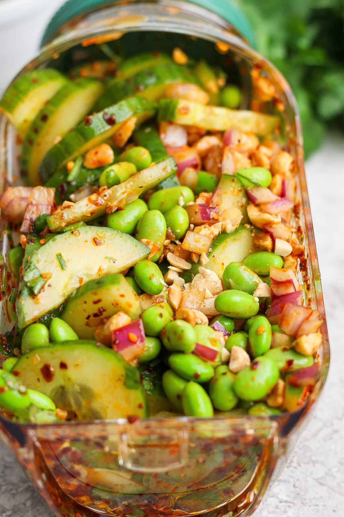 A close-up of a salad with sliced cucumbers, edamame, red onion, chopped nuts, and a red chili-flake dressing in a rectangular glass container.