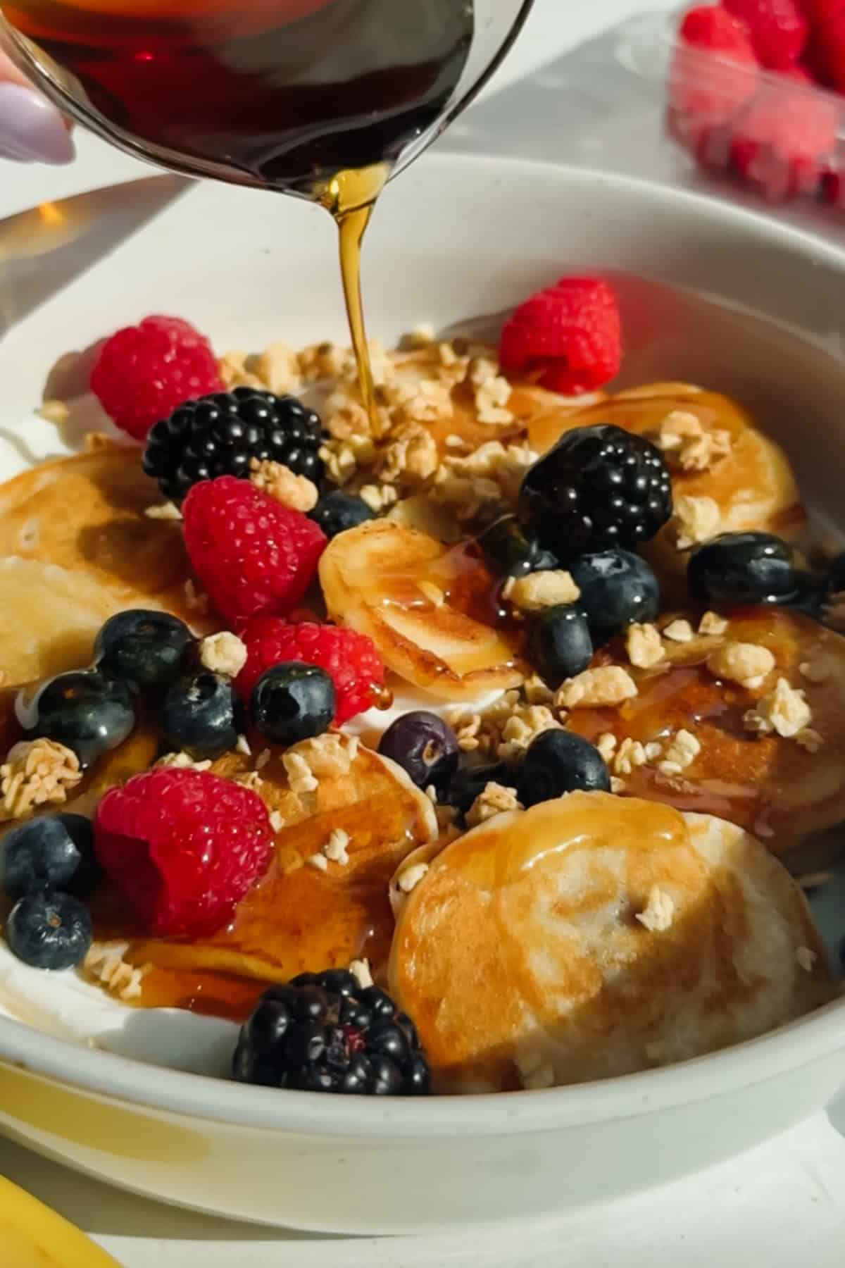 A bowl of mini pancakes topped with raspberries, blackberries, granola, and syrup being poured over them.