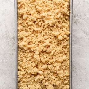 Rectangular metal baking pan filled with an even layer of unbaked, crumbly streusel or crumble topping on a light stone surface.