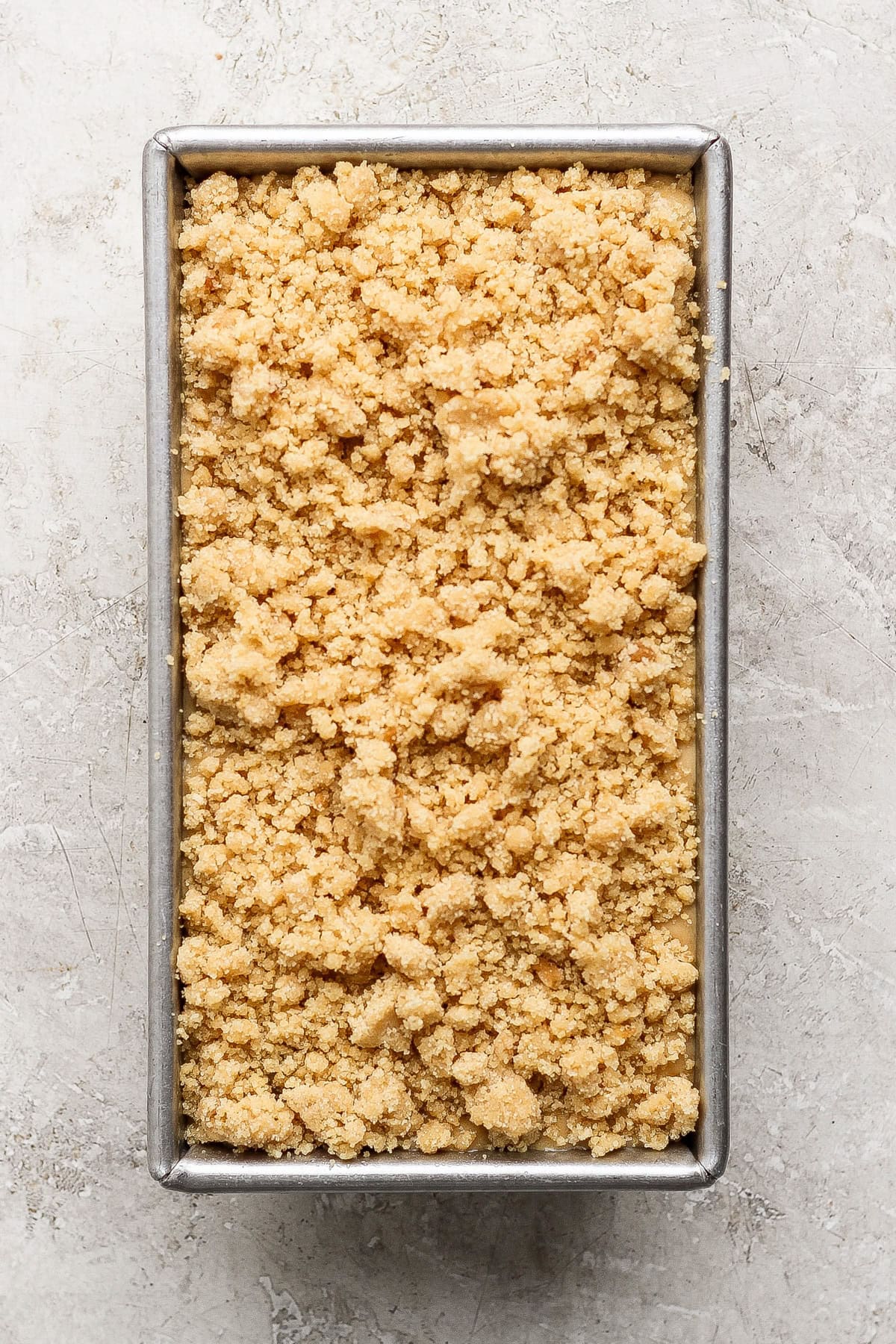 Rectangular metal baking pan filled with an even layer of unbaked, crumbly streusel or crumble topping on a light stone surface.