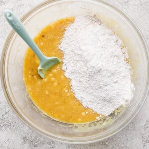 A glass bowl containing a yellow batter with a pile of white flour on top and a light blue spatula resting inside.