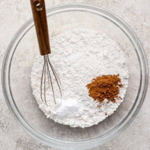 A glass bowl with flour, baking powder, baking soda, and ground cinnamon, with a metal whisk resting inside, on a light-colored surface.
