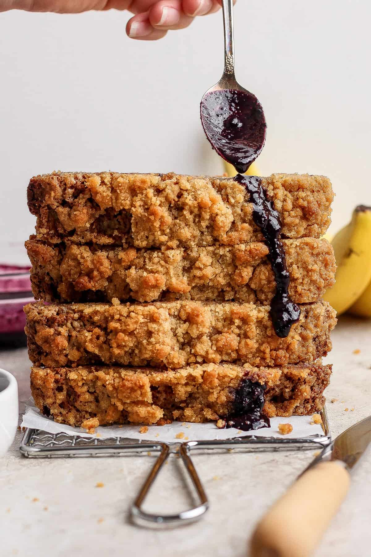 Four slices of crumb-topped bread are stacked on a wire rack as a hand drizzles dark syrup or sauce from a spoon onto the stack. Bananas are visible in the background.