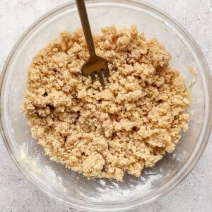 A glass bowl filled with crumbly dough mixture being mixed with a fork on a light textured surface.
