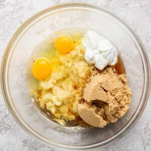 A glass bowl containing two eggs, mashed bananas, brown sugar, yogurt, and vanilla extract on a light textured surface.