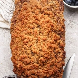 A loaf of crumb-topped bread sits on a light surface next to a knife, a bowl of blueberries, a striped cloth, and a small bowl of dark fruit jam.