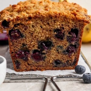 A close-up of a sliced loaf of blueberry banana bread with a crumb topping, showing blueberries inside the bread.