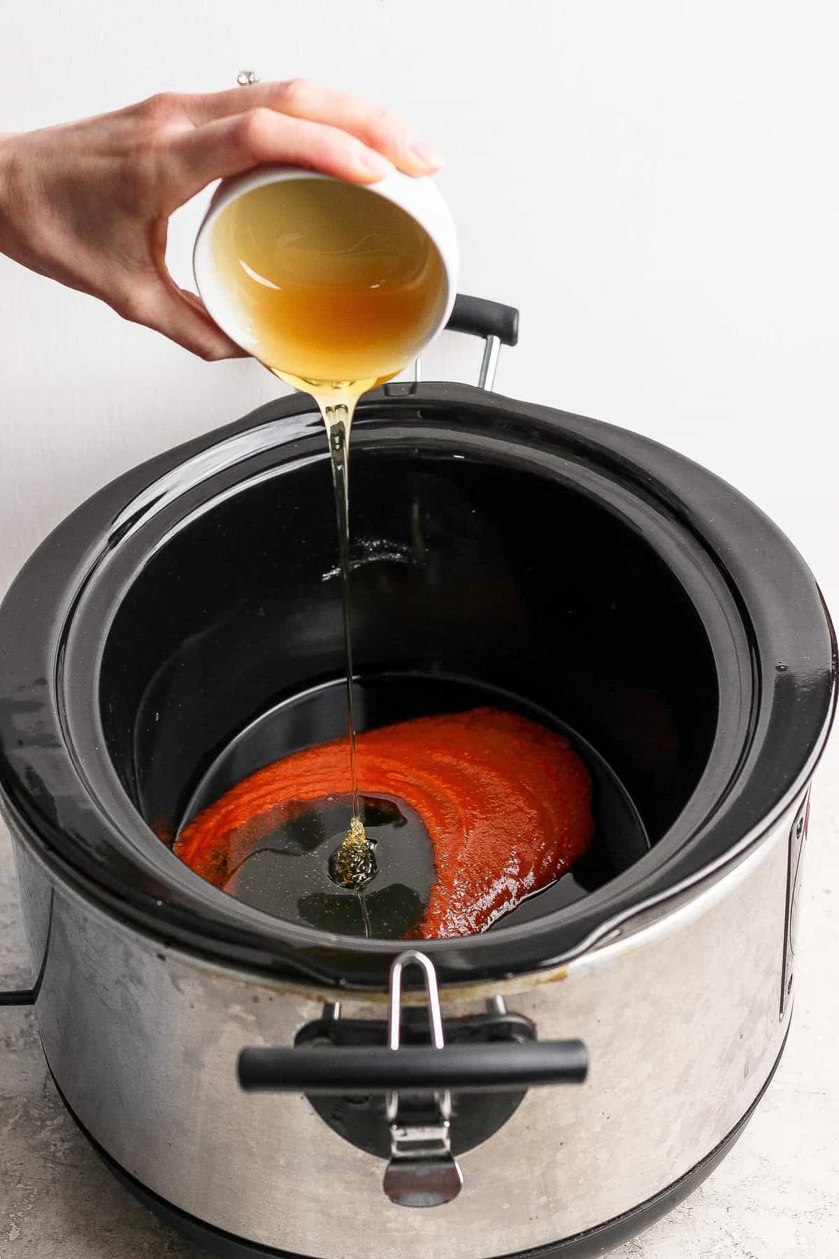 A hand pours a light brown liquid from a cup into a slow cooker containing a red sauce.