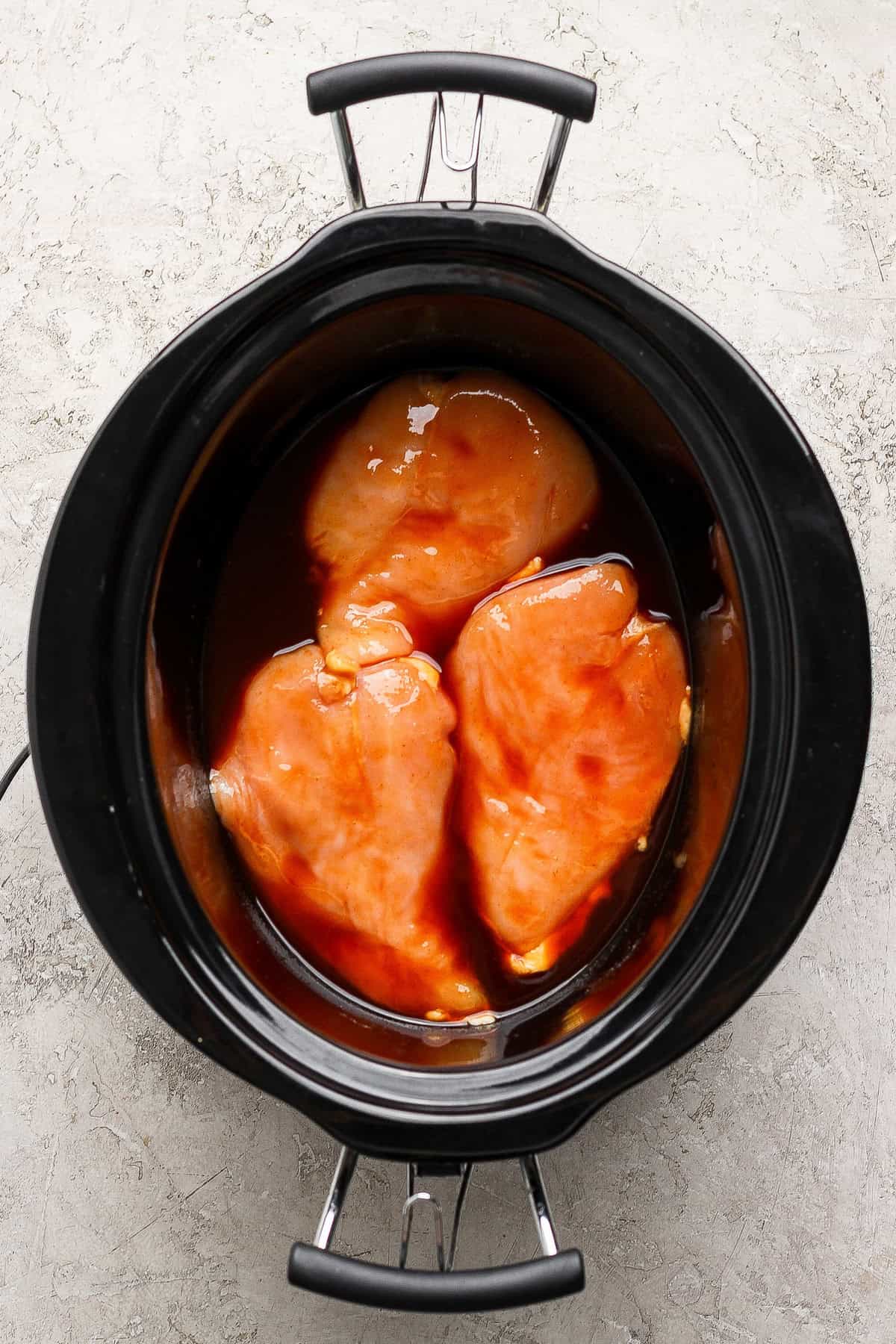 Three raw chicken breasts coated in sauce are placed in a black slow cooker on a light textured surface.