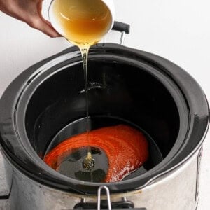 A hand pours a light brown liquid from a cup into a slow cooker containing a red sauce.