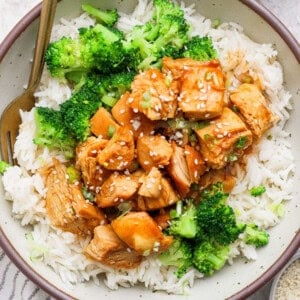 A bowl of white rice topped with cooked chicken pieces in sauce, broccoli florets, and a sprinkle of sesame seeds, with a fork on the side.