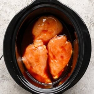 Three raw chicken breasts coated in sauce are placed in a black slow cooker on a light textured surface.