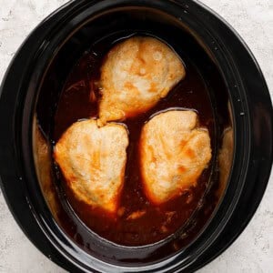 Three pieces of chicken breast cooking in a slow cooker with a dark brown sauce.