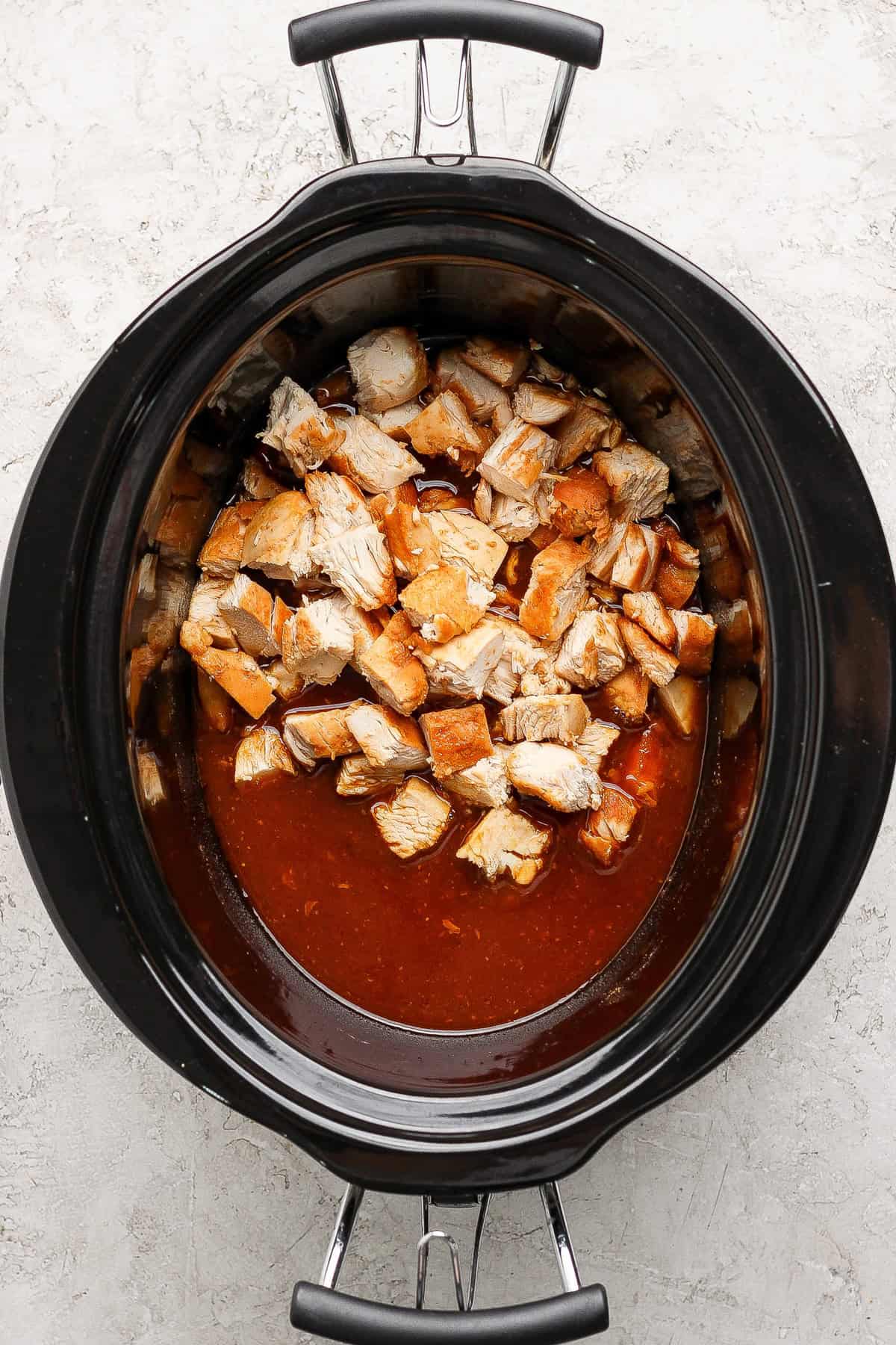 Diced cooked chicken in a slow cooker with a reddish-brown sauce, viewed from above on a light-colored surface.