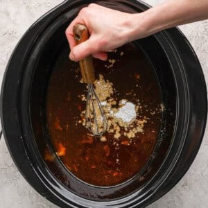 A hand uses a whisk to mix ingredients, including liquids and powders, in a black slow cooker on a light countertop.