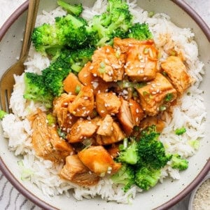 A bowl of white rice topped with broccoli and chunks of sauced chicken, garnished with sesame seeds, with a fork resting in the bowl.