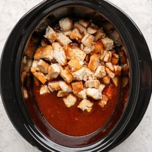 Diced cooked chicken in a slow cooker with a reddish-brown sauce, viewed from above on a light-colored surface.