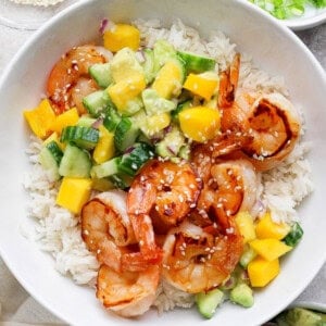 A bowl of white rice topped with grilled shrimp, diced mango, cucumber, avocado, and sprinkled with sesame seeds.