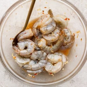 Raw shrimp marinating in a clear glass bowl with a metal spoon, mixed with a sauce containing red pepper flakes and minced garlic, on a light textured surface.