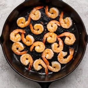 Cooked shrimp arranged in a single layer around the edge of a black cast iron skillet on a light textured surface.