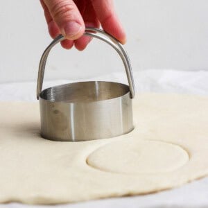 A hand presses a round metal biscuit cutter into rolled-out dough on a flat surface.