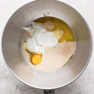A mixing bowl containing yogurt, one egg, melted butter, and liquid yeast mixture on a light textured surface.