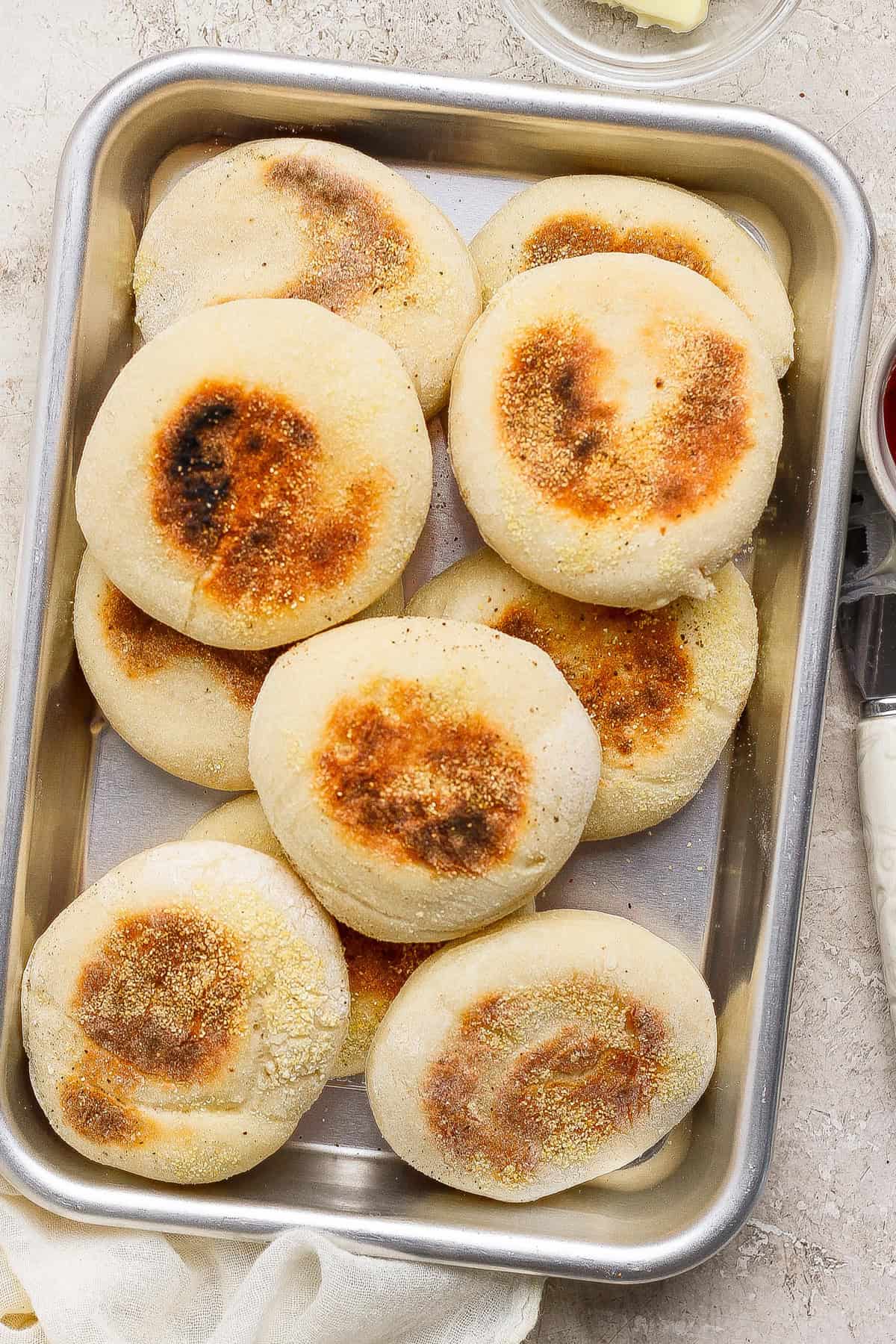 A metal tray filled with nine homemade English muffins, each with a golden-brown, toasted top and a light dusting of cornmeal.