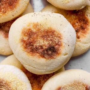 A close-up of several golden-brown English muffins topped with cornmeal, stacked together on a light surface.