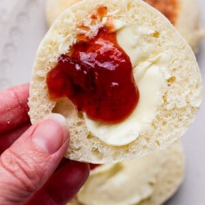 A hand holds half of an English muffin topped with melted butter and strawberry jam, with more muffins on a plate in the background.