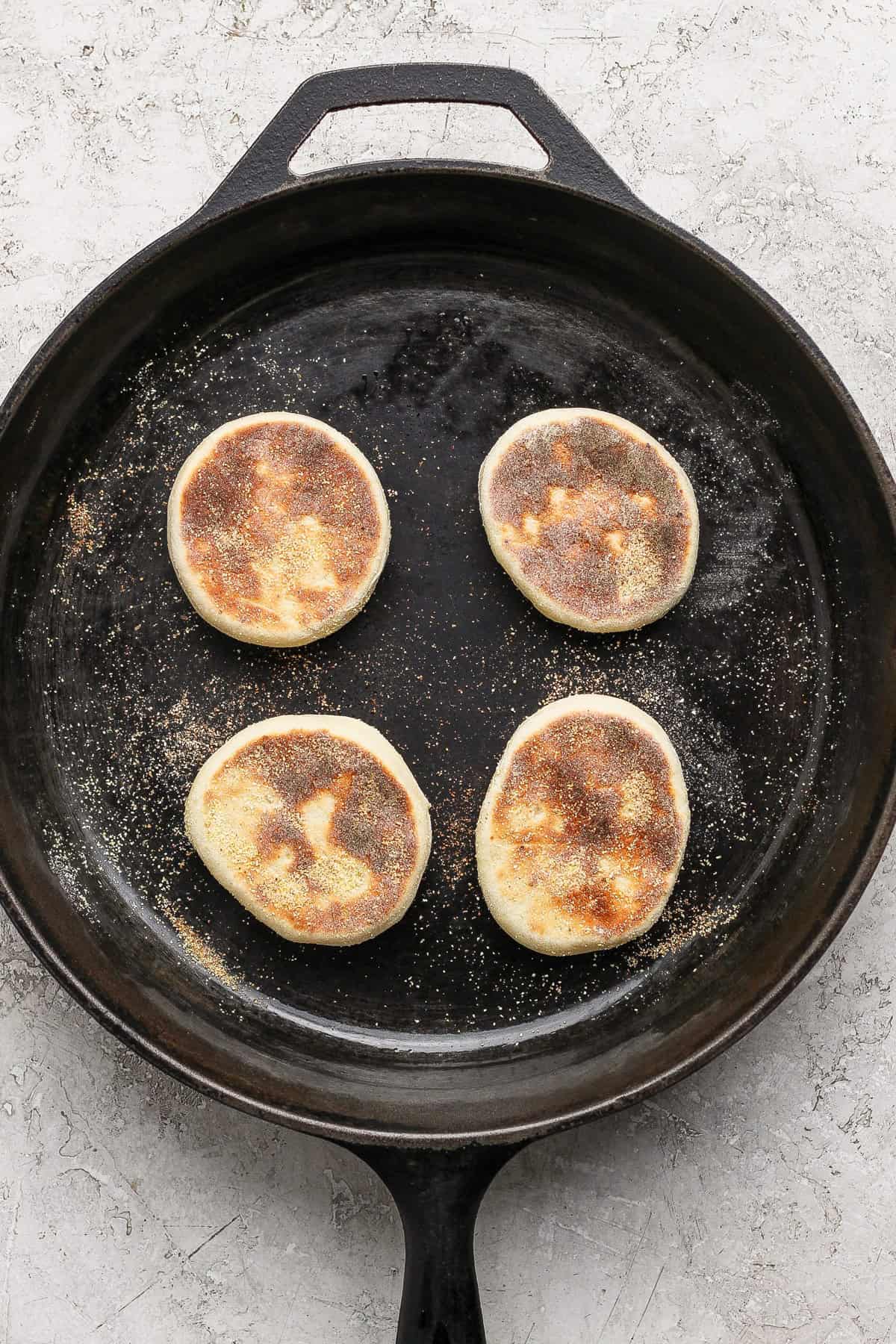 Four partially cooked English muffins in a cast iron skillet on a light textured surface.