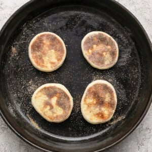 Four partially cooked English muffins in a cast iron skillet on a light textured surface.
