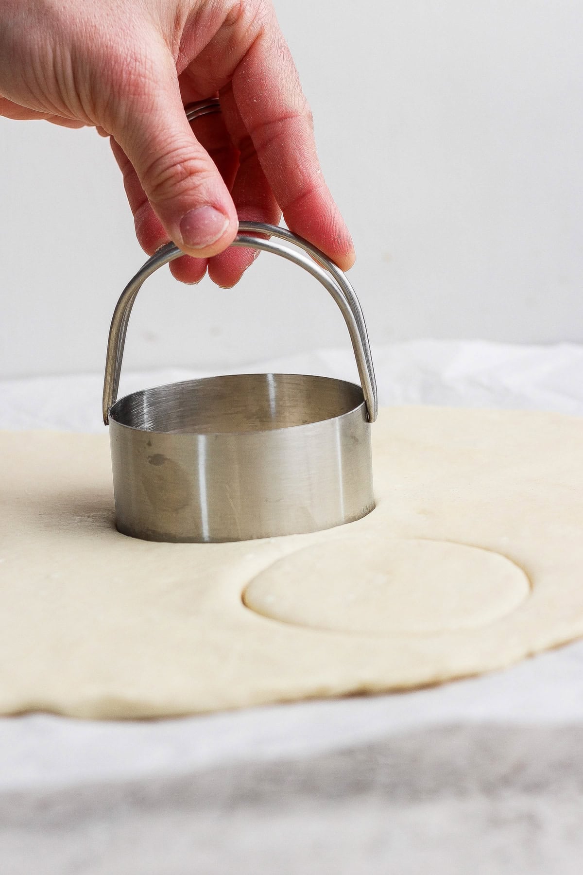 A hand presses a round metal biscuit cutter into rolled-out dough on a flat surface.