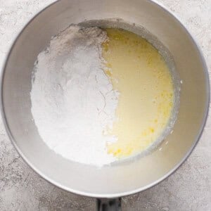 A metal mixing bowl containing flour on one side and a yellow liquid mixture on the other, placed on a light textured surface.