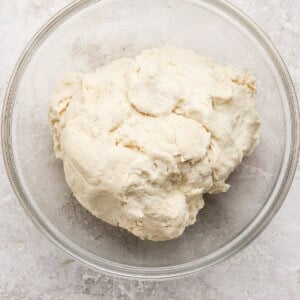 A clear glass bowl containing a ball of raw, unbaked dough sits on a light textured surface.