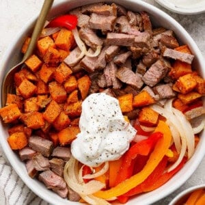 A bowl containing diced steak, roasted sweet potatoes, sautéed onions, red bell peppers, and a dollop of sour cream topped with black pepper.