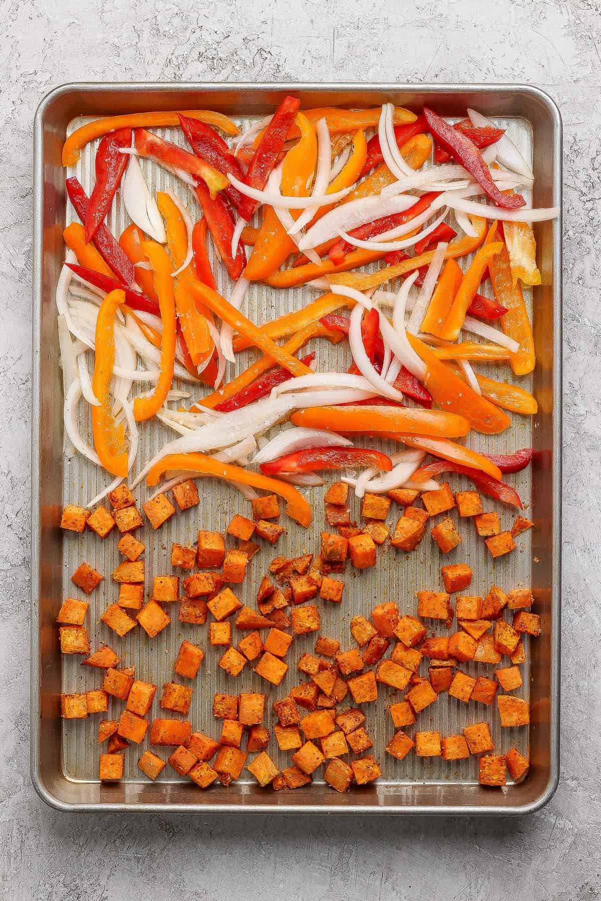 Sliced onions and bell peppers with seasoned diced sweet potatoes arranged on a baking sheet, ready for roasting.
