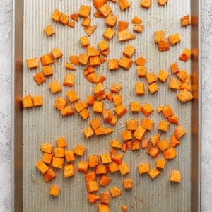 Cubed sweet potatoes seasoned with spices are spread out on a metal baking sheet, ready to be roasted in the oven.