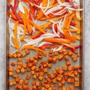 Sliced onions and bell peppers with seasoned diced sweet potatoes arranged on a baking sheet, ready for roasting.