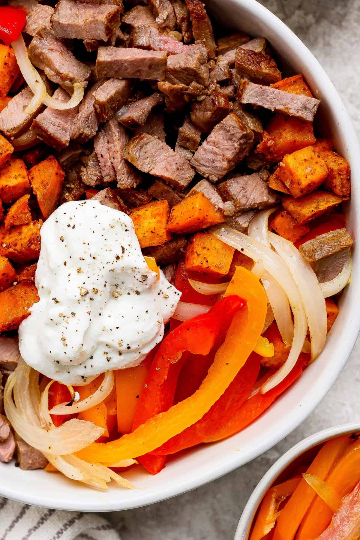 A bowl containing sliced steak, roasted sweet potatoes, sautéed onions, red and yellow bell peppers, and a dollop of sour cream, topped with cracked black pepper.