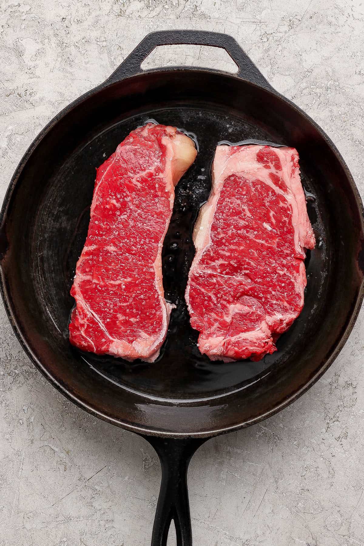Two raw steaks are placed side by side in a black cast iron skillet on a light textured surface.