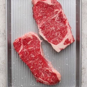 Two raw, marbled steaks sprinkled with salt are placed on a silver metal baking tray on a light-colored surface.
