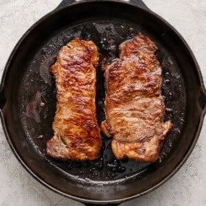 Two seared steaks are shown cooking in a black cast iron skillet on a light textured surface.