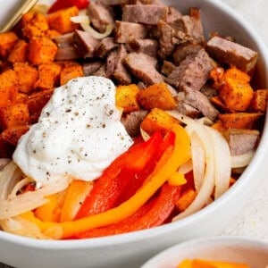 A bowl filled with sliced steak, roasted sweet potatoes, sautéed onions, bell peppers, and a dollop of sour cream, with black pepper sprinkled on top.