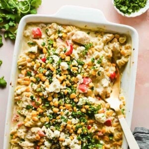 Baking dish with creamy chicken, corn, chopped red peppers, green herbs, and cheese, topped with chopped cilantro and cheese; a serving spoon is inside the dish.