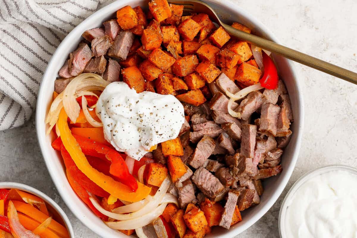 A bowl filled with diced steak, roasted sweet potatoes, sautéed onions and bell peppers, topped with a dollop of sour cream and black pepper, with a fork resting inside.