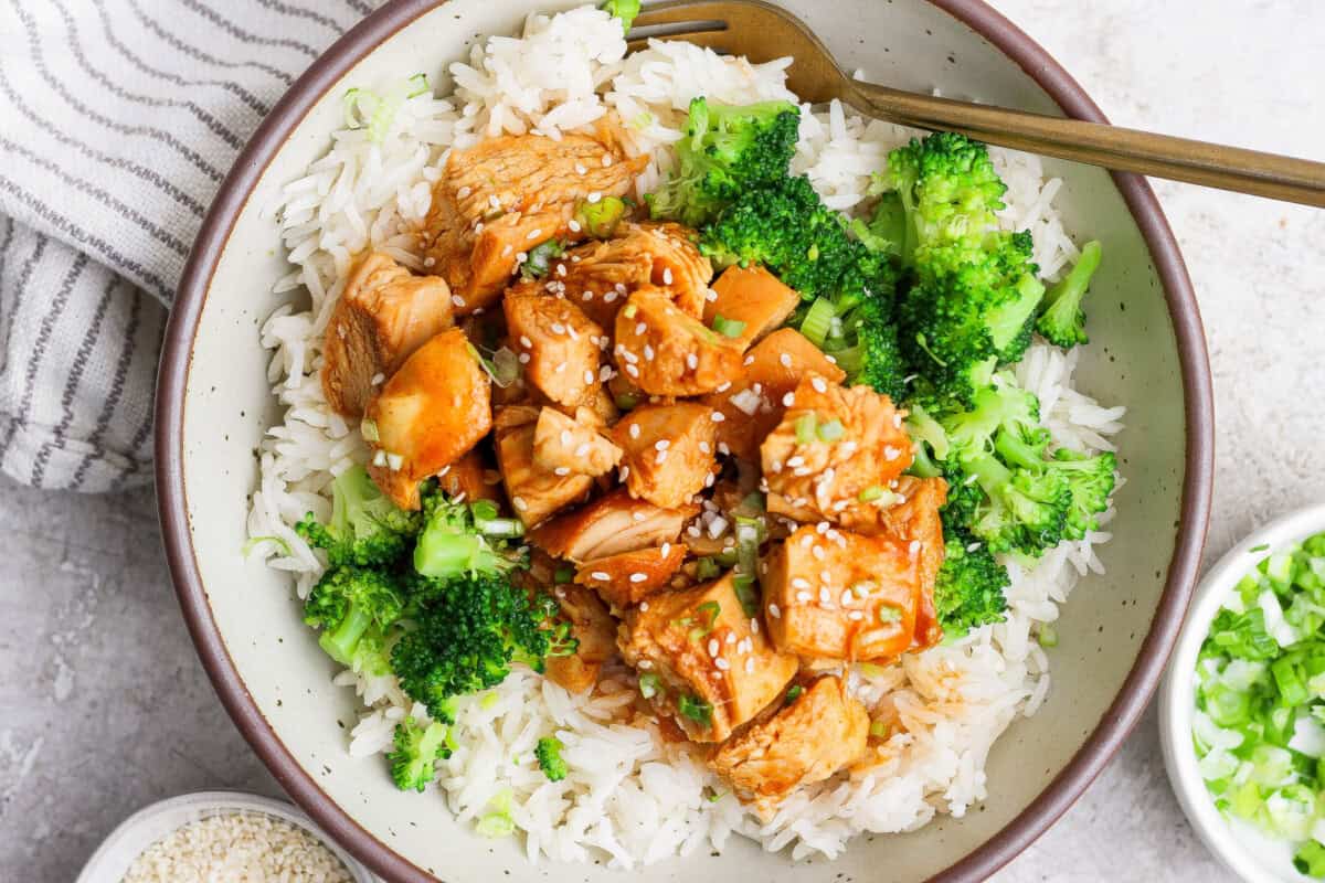 A bowl of white rice topped with broccoli, diced chicken in a brown sauce, and sprinkled with sesame seeds, with a fork on the side.